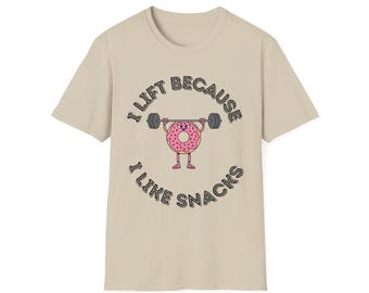 Peut inclure: T-shirt beige avec un dessin d'un beignet soulevant des poids. Le beignet rose est parsemé de vermicelles et a des bras et des jambes. Le texte autour du beignet dit "I LIFT BECAUSE I LIKE SNACKS" en noir, en forme de courbe.