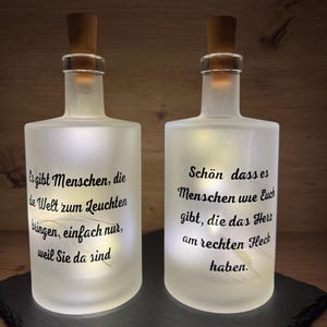 Könnte beinhalten: Zwei mattierte Glasflaschen mit Korken, von innen beleuchtet. Jede Flasche hat einen schwarzen Schriftzug. Die Flaschen stehen auf einer schwarzen Oberfläche vor einem Holzgrund. Die Flaschen sind dekorativ.