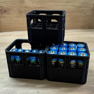 Könnte beinhalten: Drei schwarze Kunststoff-Aufbewahrungskisten, jede gefüllt mit Batterien. Zwei Kisten enthalten AA-Batterien und eine AAA-Batterien. Die Batterien haben blaue und silberne Etiketten. Die Kisten stehen auf einer Holzoberfläche.