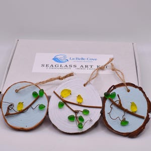 May include: Three handcrafted seaglass art ornaments with a bird theme. Each ornament features a wooden slice with a painted blue or white background, a twig, yellow glass birds, and green glass leaves. The ornaments are hanging from twine.