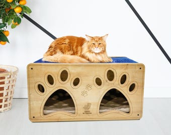 Può includere: Un letto per gatti in legno con due ingressi a forma di impronte di zampe. Il letto ha un cuscino blu in cima e un gatto è sdraiato su di esso. Il letto è in legno naturale e ha un colore marrone chiaro.