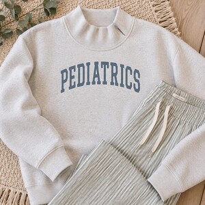 Può includere: Una felpa grigio chiaro a collo alto con la parola PEDIATRICS stampata in un carattere blu scuro arcuato sul petto, abbinata a pantaloni a righe verticali verde chiaro e bianco con una coulisse bianca in vita elastica.