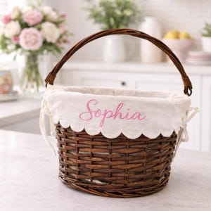 May include: A brown woven basket with a curved handle. The basket has a white scalloped liner embroidered with the name "Sophia" in pink. The basket is sitting on a white countertop.