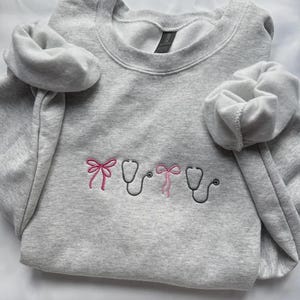 May include: A heather gray sweatshirt with embroidered medical-themed designs. The design includes two stethoscopes and two pink bows. The sweatshirt has a crew neck and long sleeves with cuffed wrists.