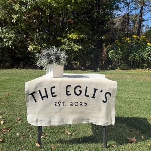 Può includere: Un tavolino con una tovaglia di lino color crema con la scritta nera "THE EGLI'S EST. 2025". Sopra, una fioriera quadrata bianca con piante verdi. Il tutto è su un prato.