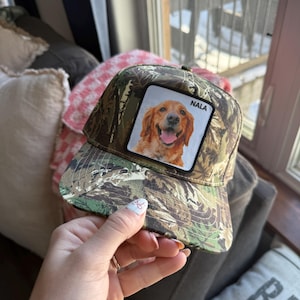 May include: Camouflage print baseball cap featuring a patch with a smiling golden retriever dog and the name "NALA". The cap has a curved brim and a black mesh back.