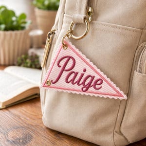 May include: A pink and white pennant-shaped keychain with the name "Paige" embroidered in a dark pink script. The keychain is attached to a beige bag with a gold-colored ring. The pennant has a scalloped edge.