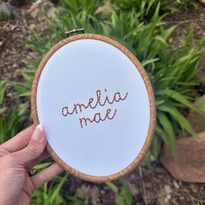 Può includere: Un telaio da ricamo rotondo con il nome "amelia mae" ricamato con filo marrone-rossastro su tessuto bianco. Il telaio ha una cornice in legno. Lo sfondo mostra un fogliame verde.