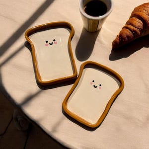Peut inclure: Deux plateaux en céramique en forme de pain de mie avec des visages souriants. Les plateaux ont un intérieur blanc et un bord brun en forme de croûte. Une tasse de café et un croissant sont en arrière-plan, suggérant un thème de petit-déjeuner.