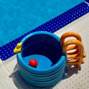 Könnte beinhalten: Ein blauer Keramikbecher in Form eines Swimmingpools mit horizontalen Rillen an der Außenseite. Der Becher hat einen orangefarbenen leiterförmigen Henkel an der Seite, eine kleine gelbe Ente am Rand und einen winzigen roten Ball am Boden.