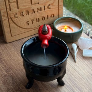 May include: A black ceramic cauldron incense burner with three legs and a red attachment at the rim holding a lit incense cone. White smoke flows downward from the cone into the cauldron. A wooden sign reads HEL CERAMIC STUDIO.
