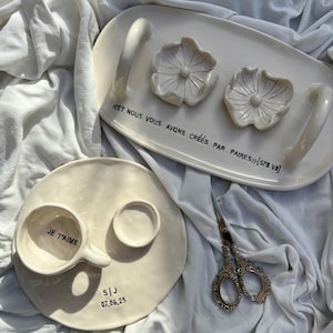 May include: Off-white ceramic serving tray with two flower-shaped dishes and handles. A matching plate with a cup that reads "JE T'AIME" and a small dish. Decorative scissors are also present. The tray has text that reads "ET NOUS VOUS AVONS CRÉES PAR PAIRES".