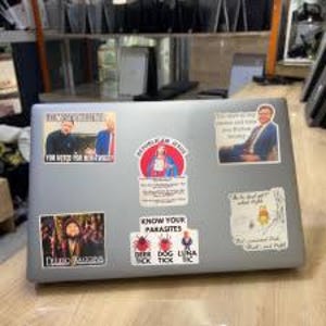 May include: Silver laptop covered in stickers. Stickers include images of people, text that reads "Know Your Parasites", and other graphics. The laptop is on a light wood surface.