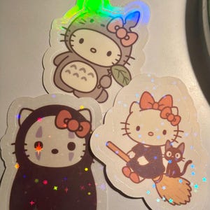 May include: Three holographic stickers featuring Hello Kitty in various costumes. One sticker shows Hello Kitty dressed as Totoro, another as No-Face, and the last as a witch with a black cat on a broom.