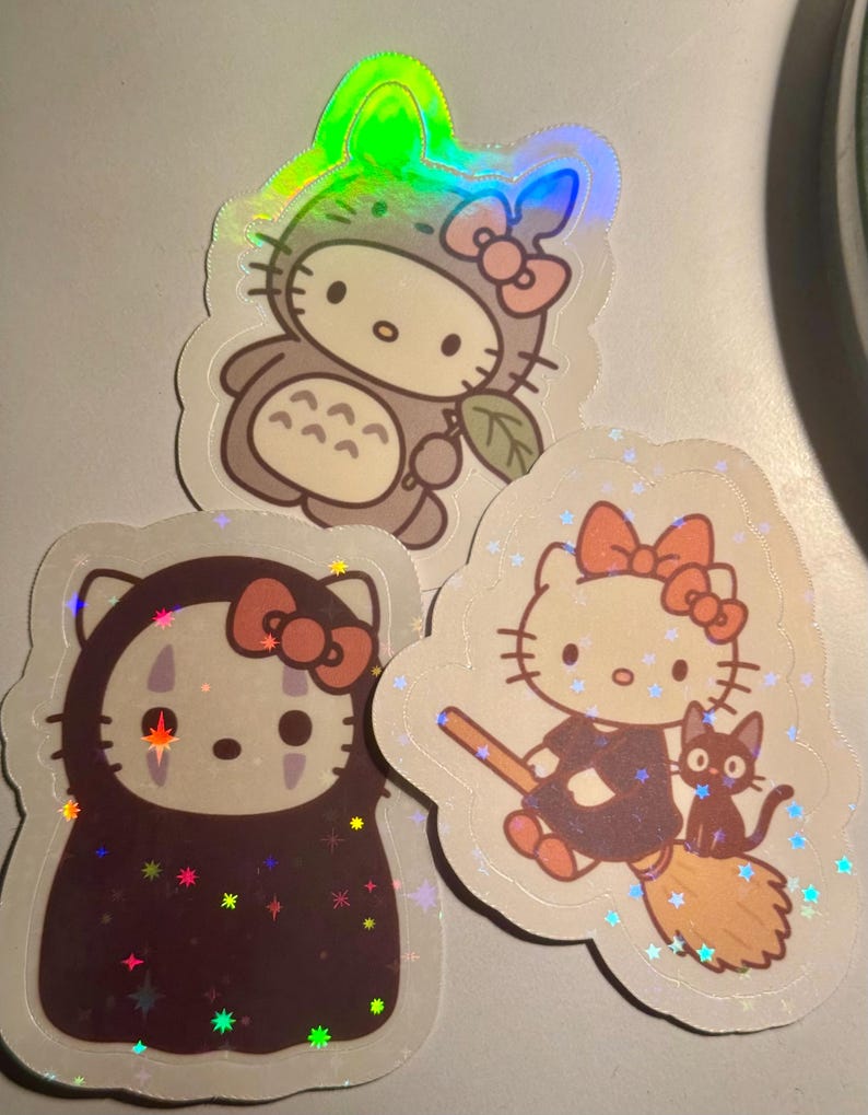 May include: Three holographic stickers featuring Hello Kitty in various costumes. One sticker shows Hello Kitty dressed as Totoro, another as No-Face, and the last as a witch with a black cat on a broom.