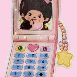 May include: A pink sticker in the shape of a flip phone featuring a cartoon character with dark hair and a pink outfit. The phone has a keypad with blue buttons and a heart-shaped button. A yellow star charm hangs from the phone.