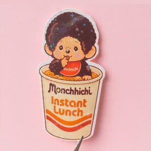 May include: A sticker featuring a Monchhichi character in a cup of instant noodles. The Monchhichi is brown with a red bib that says "onchhichi". The cup is beige with orange and red stripes and the words "Monchhichi Instant Lunch".