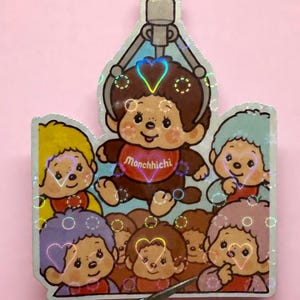 May include: A holographic sticker featuring a Monchhichi character being lifted by a claw machine. The sticker has a pink background and includes several other Monchhichi characters with heart-shaped outlines. The Monchhichi character in the claw machine has a red heart-shaped shirt with the word "Monchhichi".