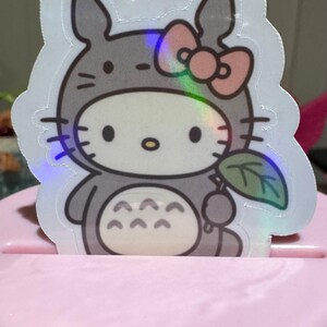 May include: A holographic sticker featuring a cartoon character in a gray Totoro costume with a pink bow. The sticker has a white border and is holding a green leaf. The sticker is placed on a pink surface.