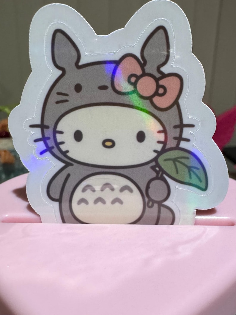 May include: A holographic sticker featuring a cartoon character in a gray Totoro costume with a pink bow. The sticker has a white border and is holding a green leaf. The sticker is placed on a pink surface.
