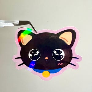 May include: A holographic sticker of a black cat with large, sparkling eyes. The cat has a pink outline, tan ears, and a blue collar with a yellow button. The sticker is held by tweezers.