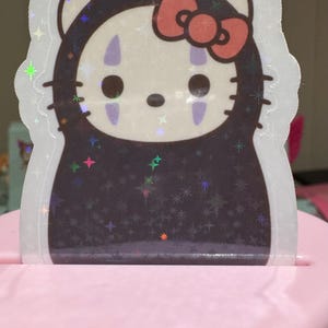 May include: A holographic sticker featuring a mashup of Hello Kitty and the character No-Face from Spirited Away. The sticker has a white outline and a pink bow. The background is black with star-shaped sparkles.