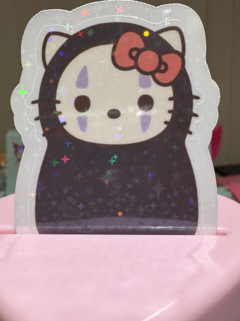 May include: A holographic sticker featuring a mashup of Hello Kitty and the character No-Face from Spirited Away. The sticker has a white outline and a pink bow. The background is black with star-shaped sparkles.