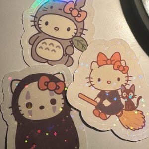 May include: Three holographic stickers featuring Hello Kitty in various costumes. One sticker shows Hello Kitty dressed as Totoro, another as No-Face, and the third as a witch with a black cat. Each sticker has a pink bow.