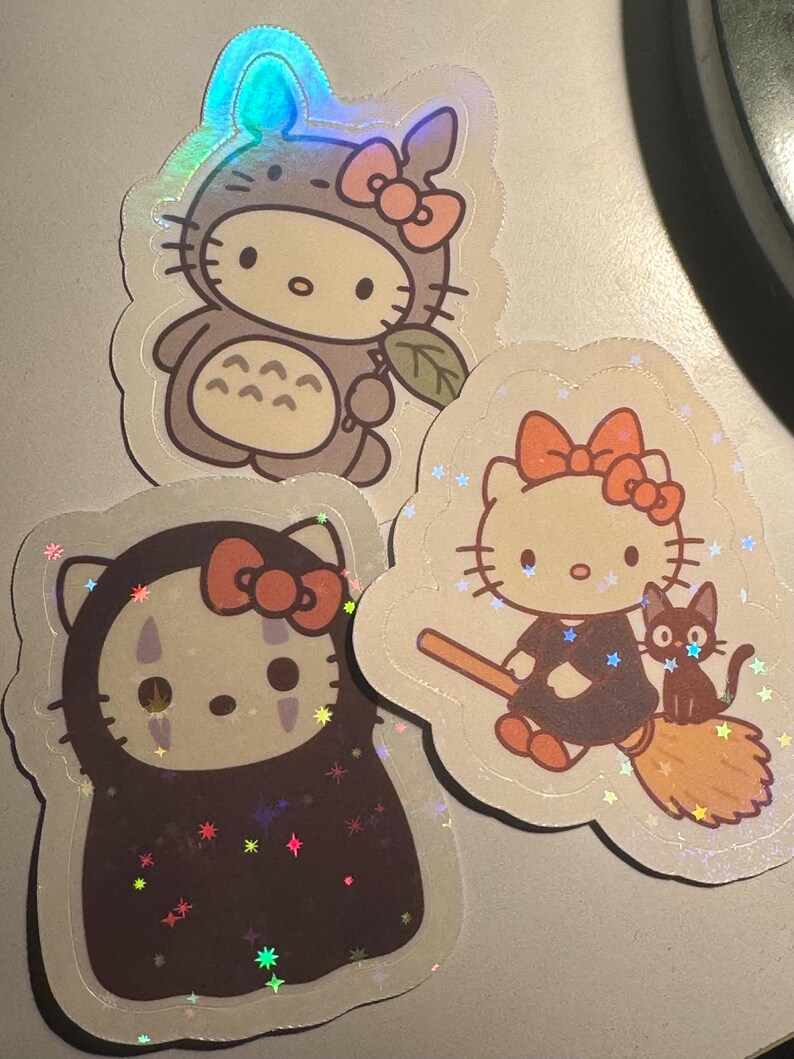 May include: Three holographic stickers featuring Hello Kitty in various costumes. One sticker shows Hello Kitty dressed as Totoro, another as No-Face, and the third as a witch with a black cat. Each sticker has a pink bow.