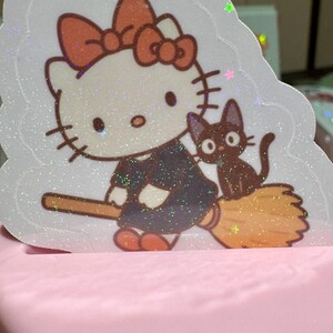May include: A glittery sticker featuring a cartoon cat character with a large bow, dressed in a dark outfit, riding a broom with a black cat companion. The sticker has a white border and is set against a pink background.