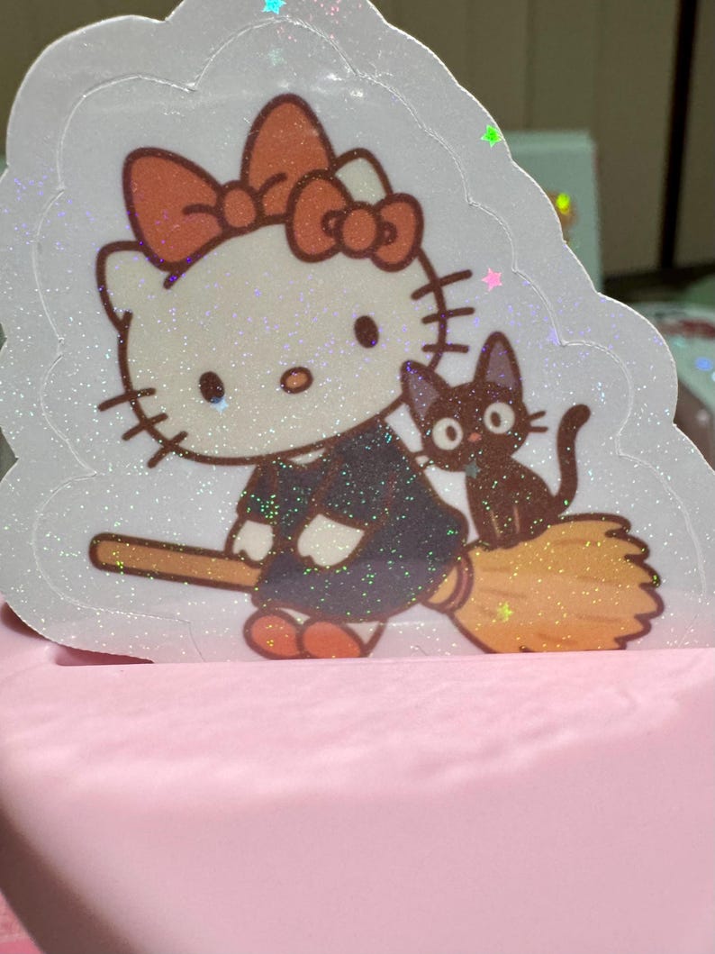 May include: A glittery sticker featuring a cartoon cat character with a large bow, dressed in a dark outfit, riding a broom with a black cat companion. The sticker has a white border and is set against a pink background.
