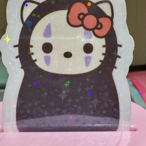 May include: A holographic sticker featuring a mashup of Hello Kitty and No-Face from Spirited Away. The sticker has a white outline, a pink bow, and a dark brown body. The design includes star-shaped sparkles.