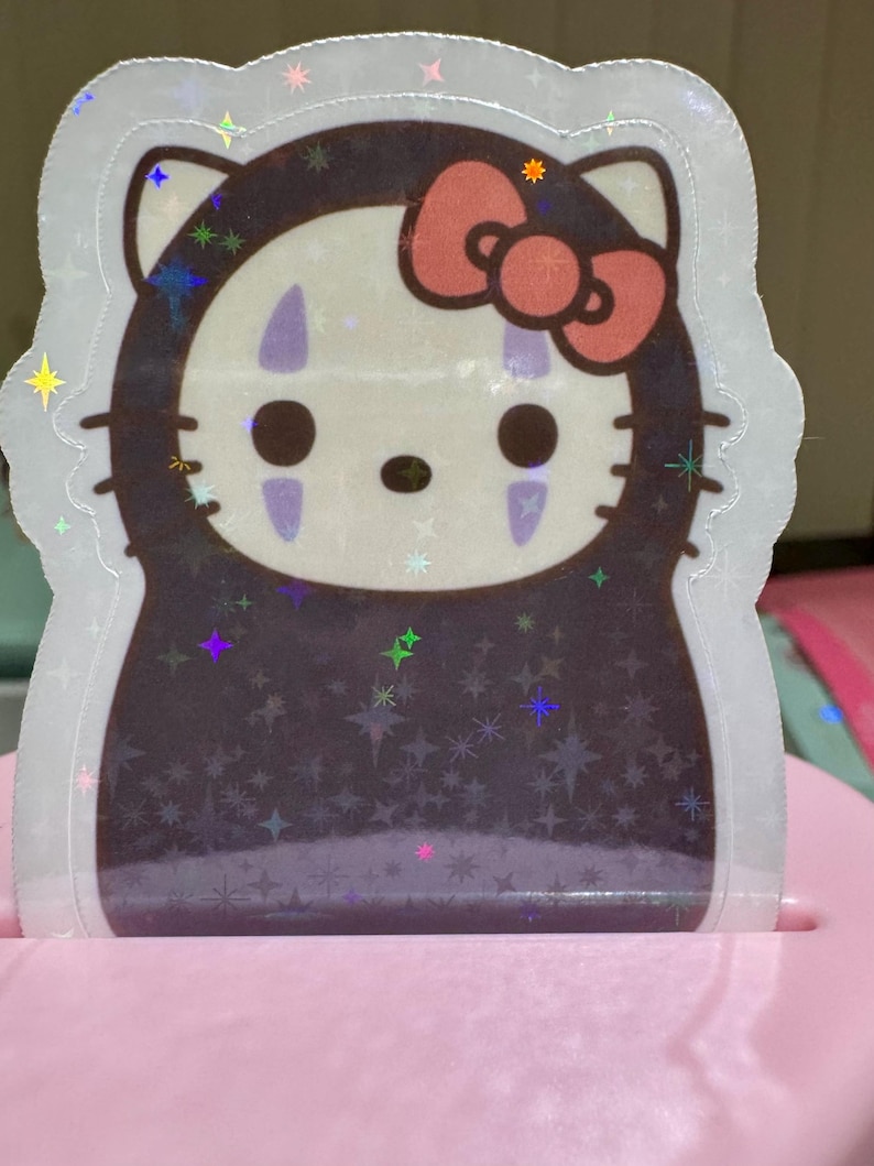 May include: A holographic sticker featuring a mashup of Hello Kitty and No-Face from Spirited Away. The sticker has a white outline, a pink bow, and a dark brown body. The design includes star-shaped sparkles.