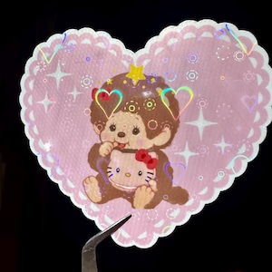 May include: A heart-shaped sticker with a pink background and a scalloped edge. The sticker features a brown cartoon monkey holding a Hello Kitty doll. The design includes holographic accents and star and heart shapes.