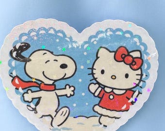 May include: A heart-shaped sticker featuring Snoopy wearing a red scarf and Hello Kitty in a red dress with a bow. The sticker has a blue background with a glittery border and is held by a metal tweezer.