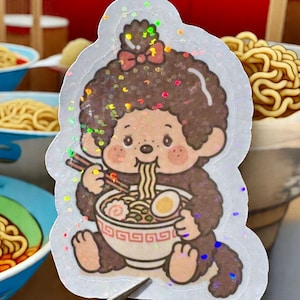 May include: A holographic sticker featuring a cartoon character eating ramen. The character is brown with a bow and is holding chopsticks and a bowl of ramen. The sticker has a glittery effect and is surrounded by a white border.