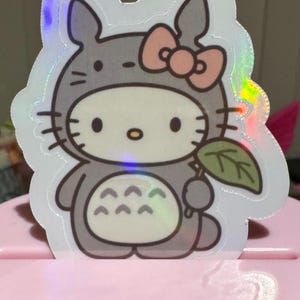 May include: A holographic sticker featuring a hybrid design of Hello Kitty and Totoro. The sticker shows Hello Kitty in a Totoro costume, holding a leaf. The design includes a pink bow, gray fur, and a white face.