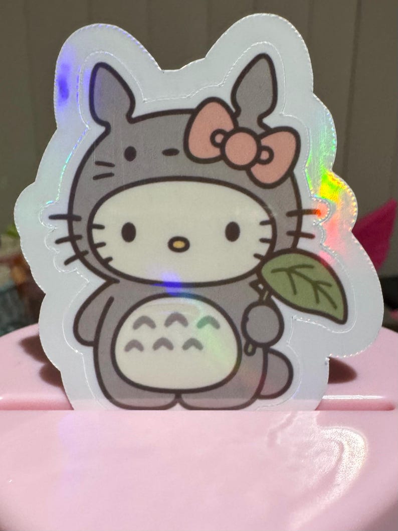 May include: A holographic sticker featuring a hybrid design of Hello Kitty and Totoro. The sticker shows Hello Kitty in a Totoro costume, holding a leaf. The design includes a pink bow, gray fur, and a white face.