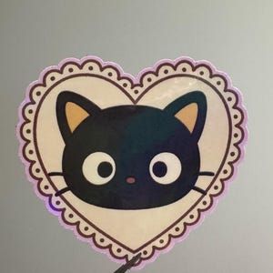 May include: A heart-shaped sticker featuring a black cat face with large white eyes and small yellow ears. The cat is centered on a cream-colored background, framed by a scalloped border in light purple and brown.