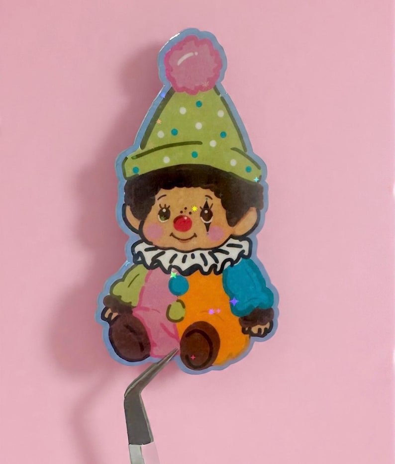 May include: A colorful clown sticker with a green polka-dot hat and a pink pom-pom. The clown has a white ruffled collar, and its outfit is a mix of pink, orange, blue, and green. The sticker is held by tweezers against a pink background.