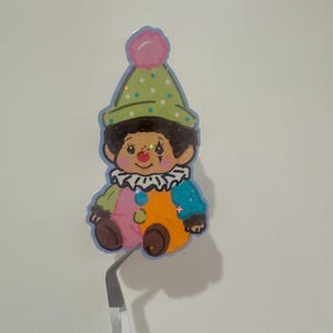 May include: A colorful sticker of a cartoon clown with a green polka-dot hat, a pink pom-pom, and a ruffled collar. The clown has a brown face, rosy cheeks, and a multi-colored outfit. The sticker is held by a pair of tweezers.