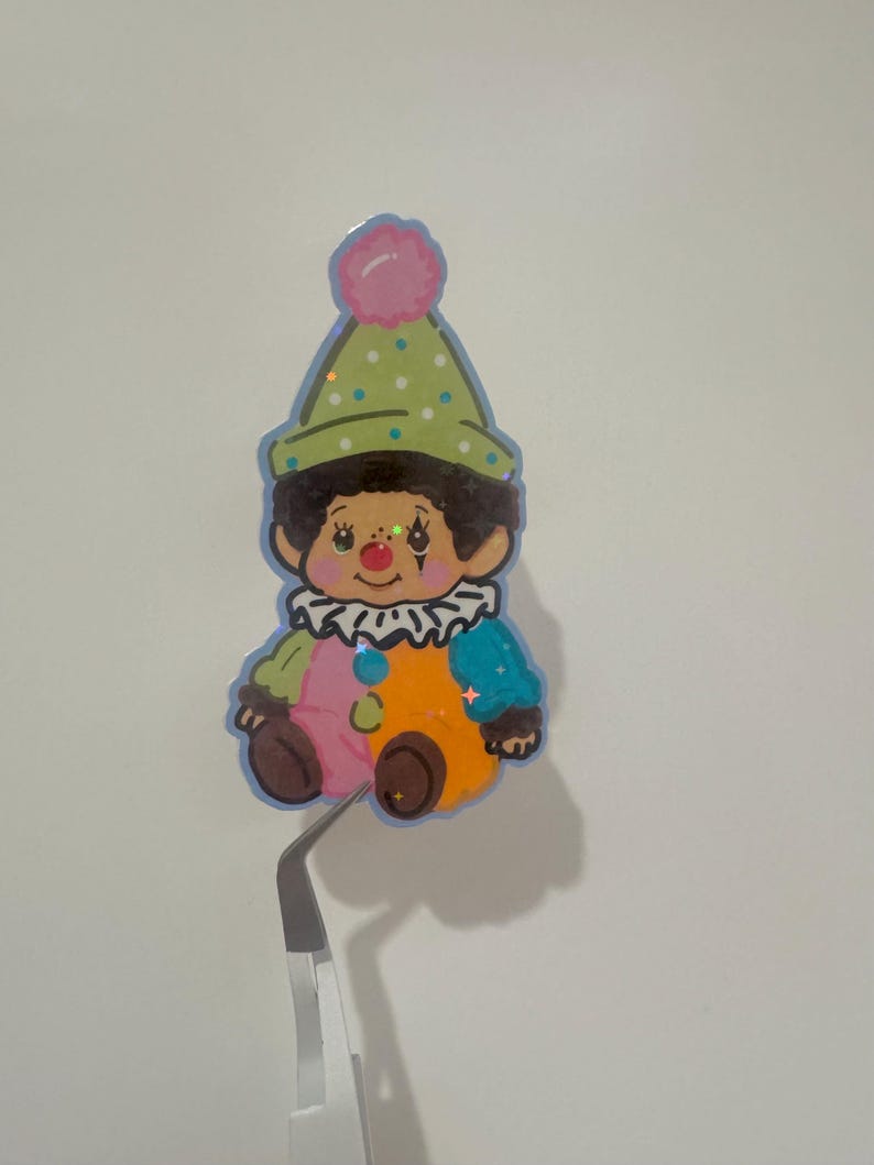 May include: A colorful sticker of a cartoon clown with a green polka-dot hat, a pink pom-pom, and a ruffled collar. The clown has a brown face, rosy cheeks, and a multi-colored outfit. The sticker is held by a pair of tweezers.