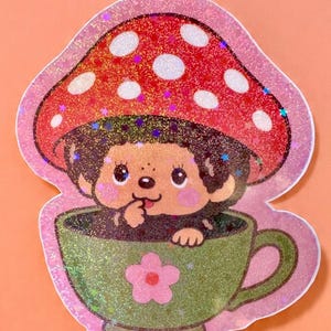May include: A whimsical sticker featuring a cartoon character with a mushroom cap hat, peeking out of a green teacup with a pink flower. The sticker has a glittery finish and a pink outline, set against an orange background.