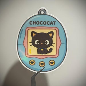 May include: A Chococat sticker in a light blue, oval-shaped design. The sticker features a black cat character with a yellow background and the word "CHOCOCAT" at the top. The sticker has a holographic effect.