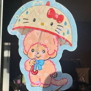 May include: A die-cut sticker featuring a cartoon dog under an umbrella. The umbrella is designed with a Hello Kitty face and a red bow. The dog is light pink with a blue bow and holding a pink umbrella. The sticker has a light blue border.