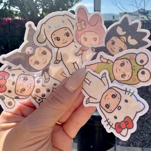 May include: A collection of colorful, die-cut stickers featuring various cartoon characters in baby-like forms. The stickers showcase characters in different outfits and poses, with a variety of colors and designs. The stickers are held in a hand.