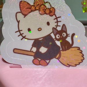May include: A holographic sticker featuring a cartoon cat character with a bow, dressed in a dark outfit, riding a broom with a black cat companion. The sticker has a glittery, iridescent effect and is set against a light pink background.