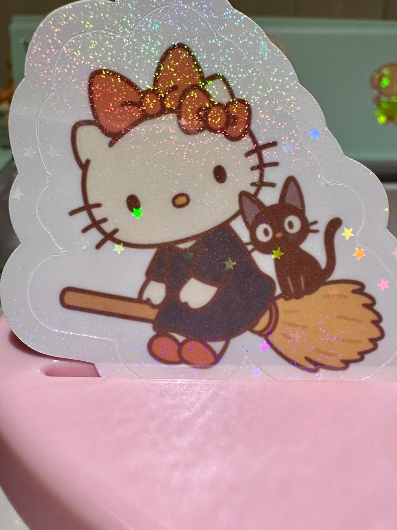 May include: A holographic sticker featuring a cartoon cat character with a bow, dressed in a dark outfit, riding a broom with a black cat companion. The sticker has a glittery, iridescent effect and is set against a light pink background.