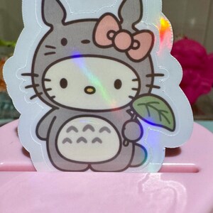 May include: A holographic sticker featuring a cartoon character mashup. The design combines Hello Kitty with a Totoro costume, complete with a pink bow, gray fur, and a leaf. The sticker has a shiny, iridescent finish.