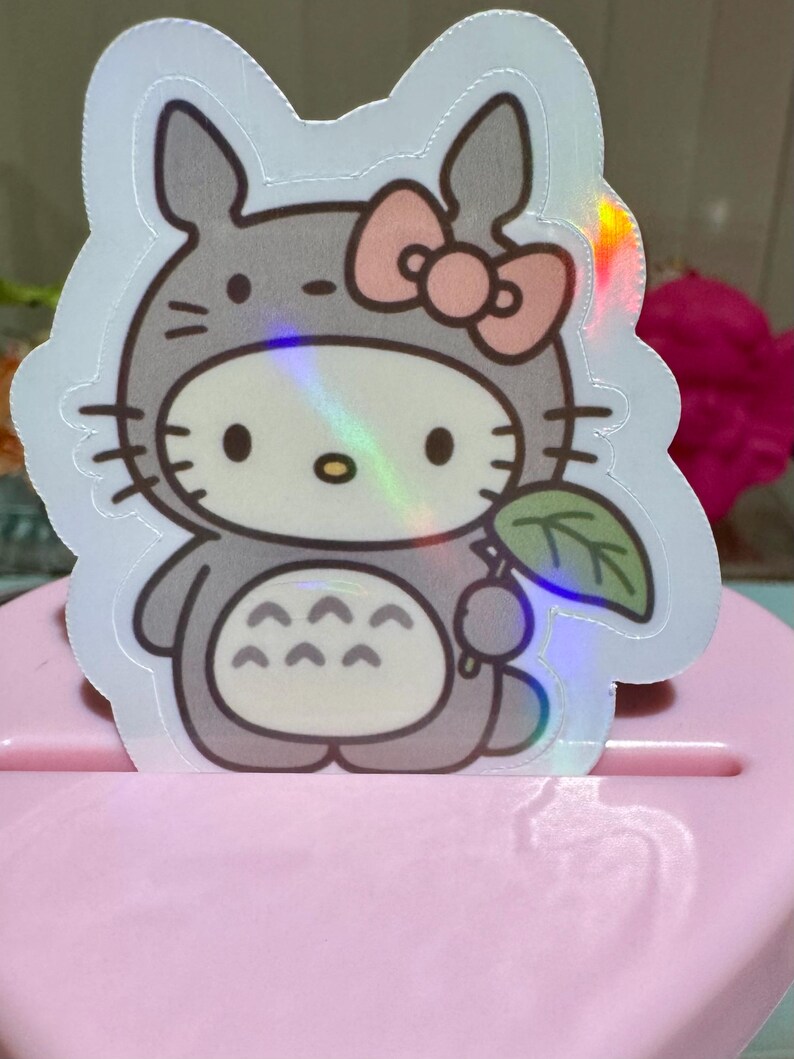 May include: A holographic sticker featuring a cartoon character mashup. The design combines Hello Kitty with a Totoro costume, complete with a pink bow, gray fur, and a leaf. The sticker has a shiny, iridescent finish.
